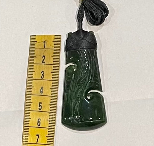 Greenstone pounamu pendant with intricate carvings on a white background, next to a ruler for scale.