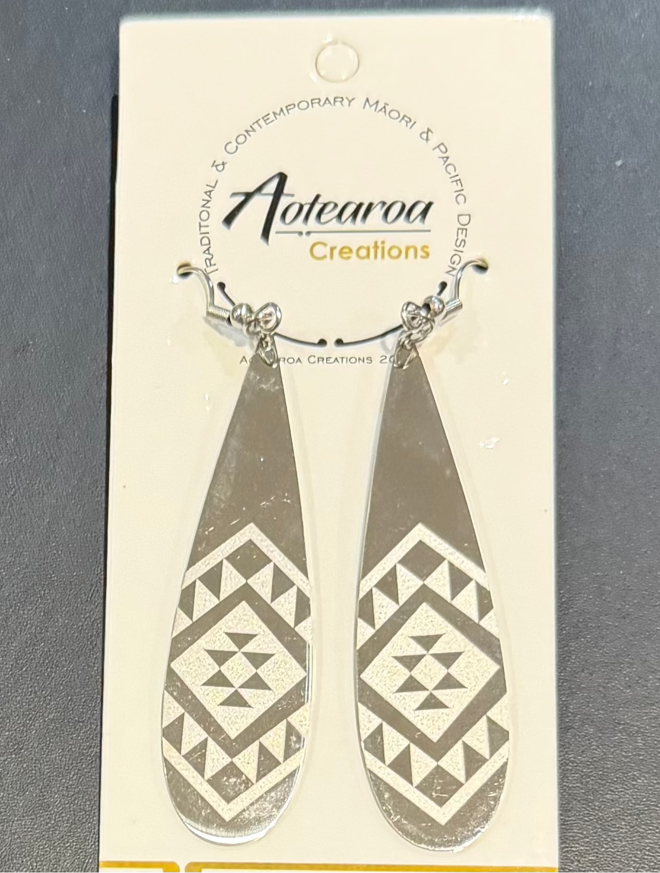 A pair of silver-toned teardrop dangle earrings with a geometric tribal pattern, hanging from a cream-colored Aotearoa Creations brand card against a dark gray background.