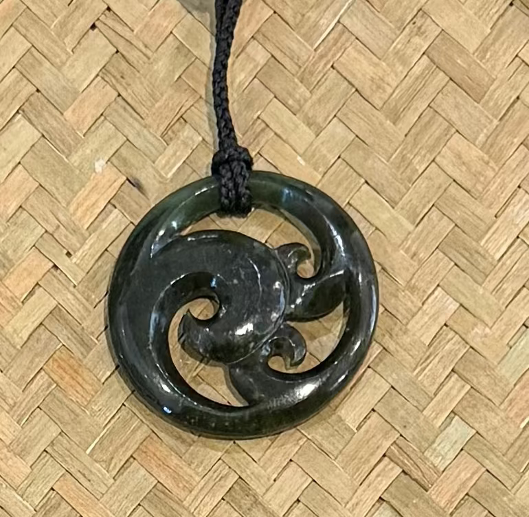 A dark green carved pendant with a swirling Polynesian-style motif, hanging from a black cord over a woven straw mat background.