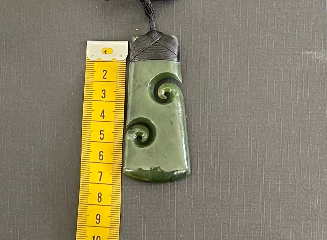 Greenstone toki jade pendant with spiral design next to a ruler on a gray background