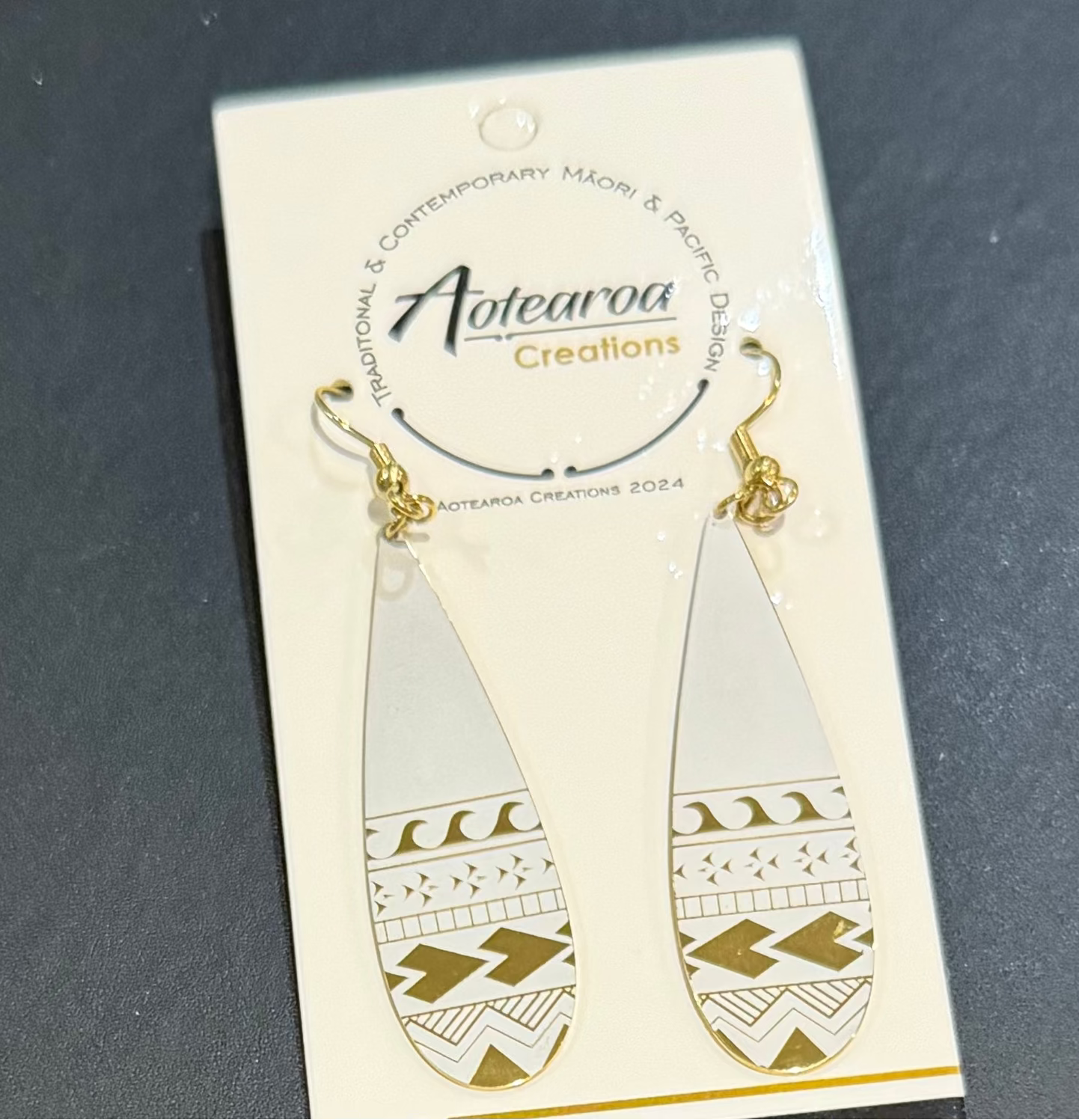 A pair of teardrop-shaped dangle earrings with gold-tone hooks and geometric patterns, hanging from a white display card labeled Aotearoa Creations.
