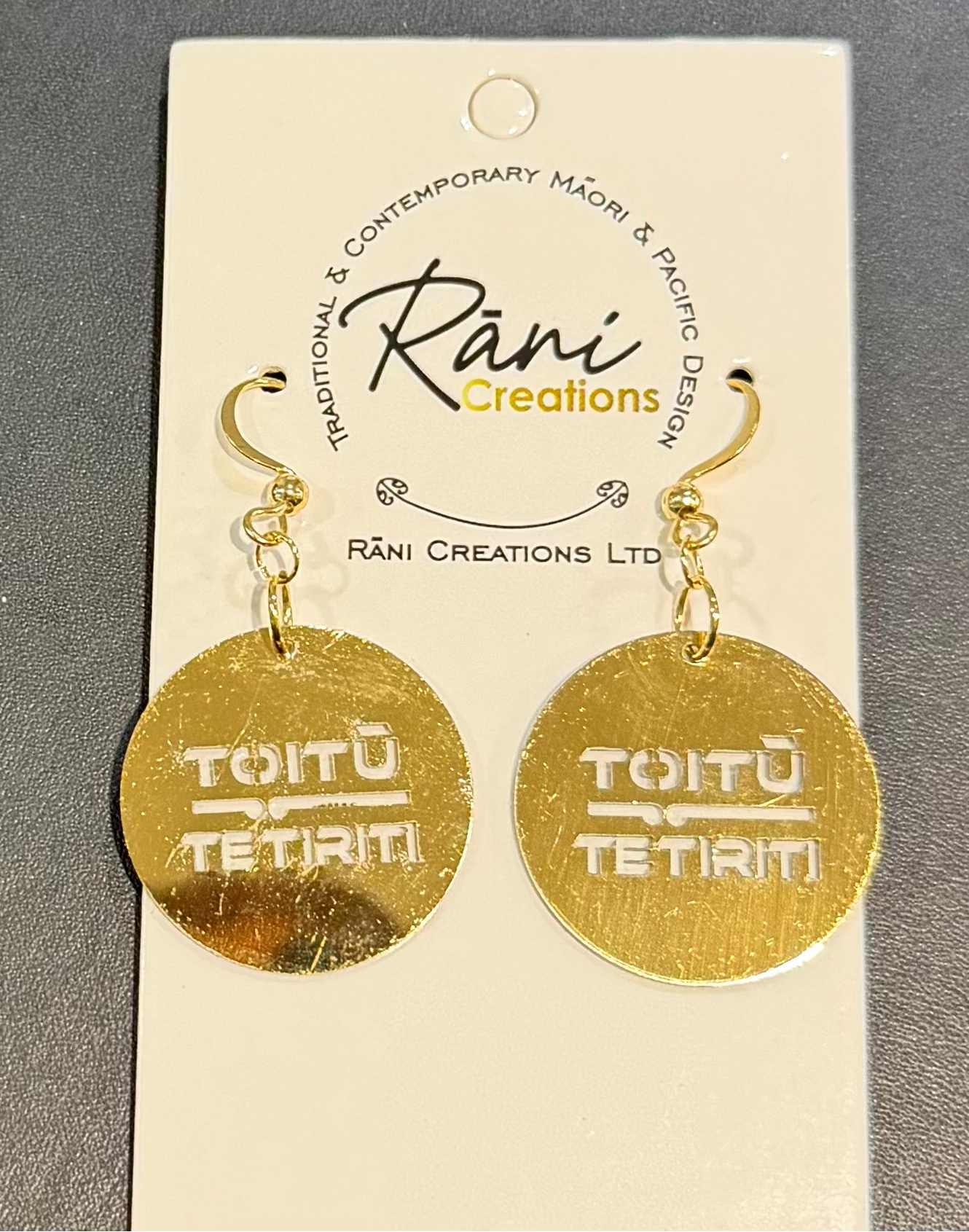 A pair of gold disc dangle earrings engraved with 'Toitu TeTiriti' hanging from gold hooks on a white brand card by Rani Creations, set against a dark gray textured background.