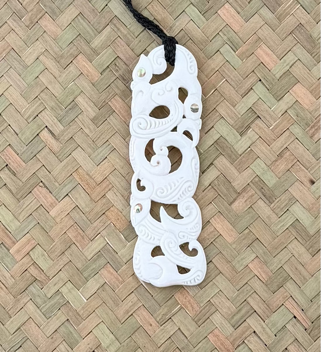 White decorative Manaia pendant on a black cord against a woven background