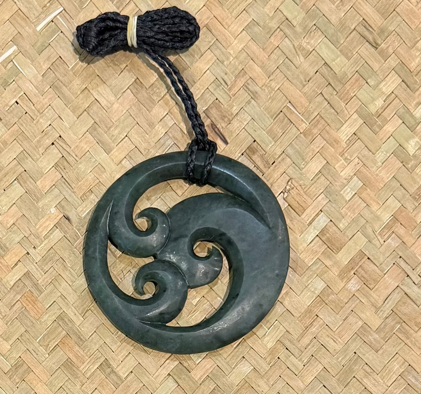 A greenstone circular pendant with a triple swirl motif, attached to a black braided cord, displayed on a woven straw mat background.