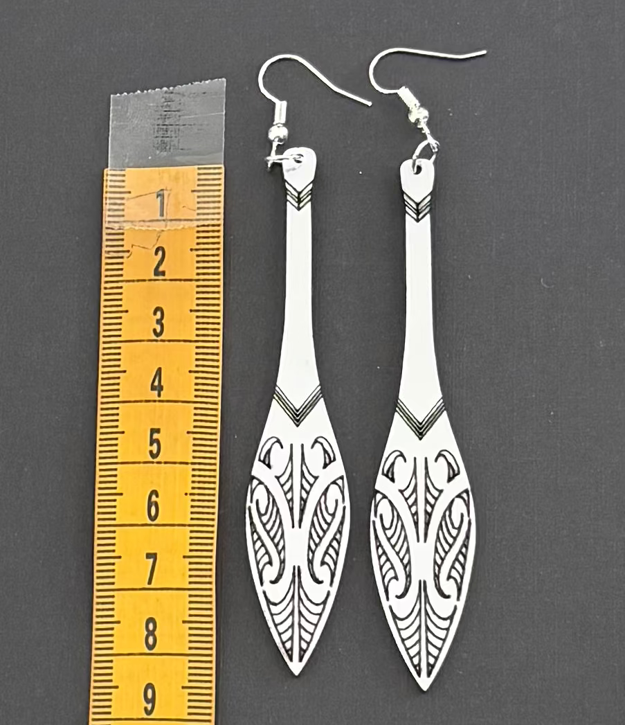 Pair of long, thin earrings with intricate designs next to a ruler on a dark background