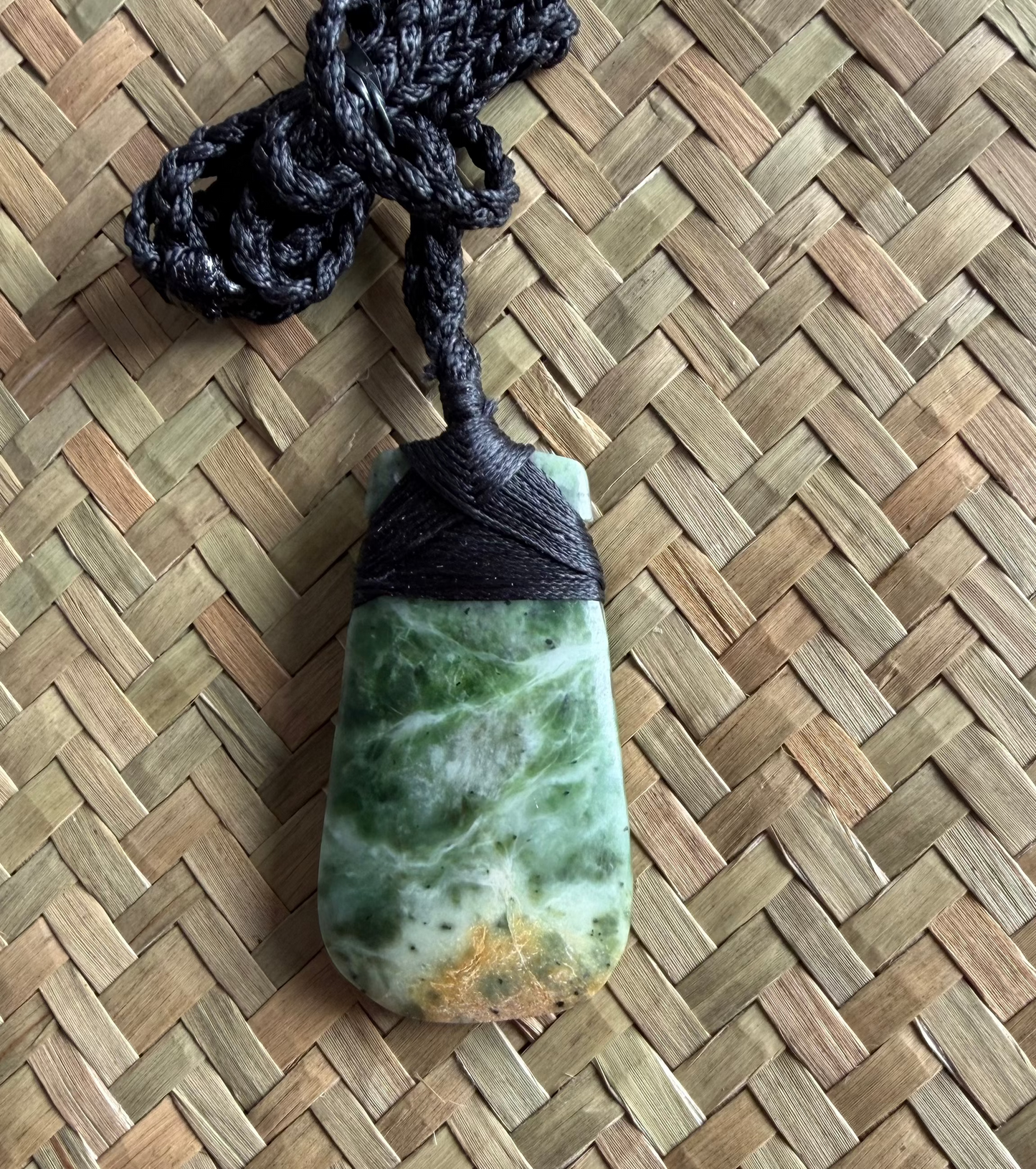 A greenstone jadeite pendant with natural marbling and a small gold spot near the bottom, wrapped with a black cord and hanging on a woven straw mat.