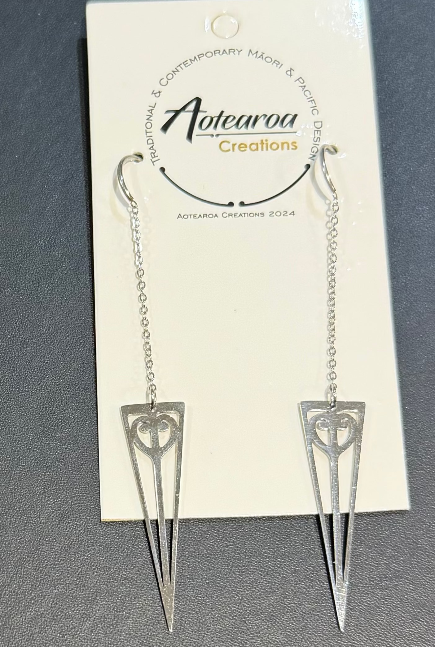 A pair of silver dangle earrings with geometric heart and arrowhead designs, hanging from a white display card labeled 'Aotearoa Creations' on a dark gray textured surface.