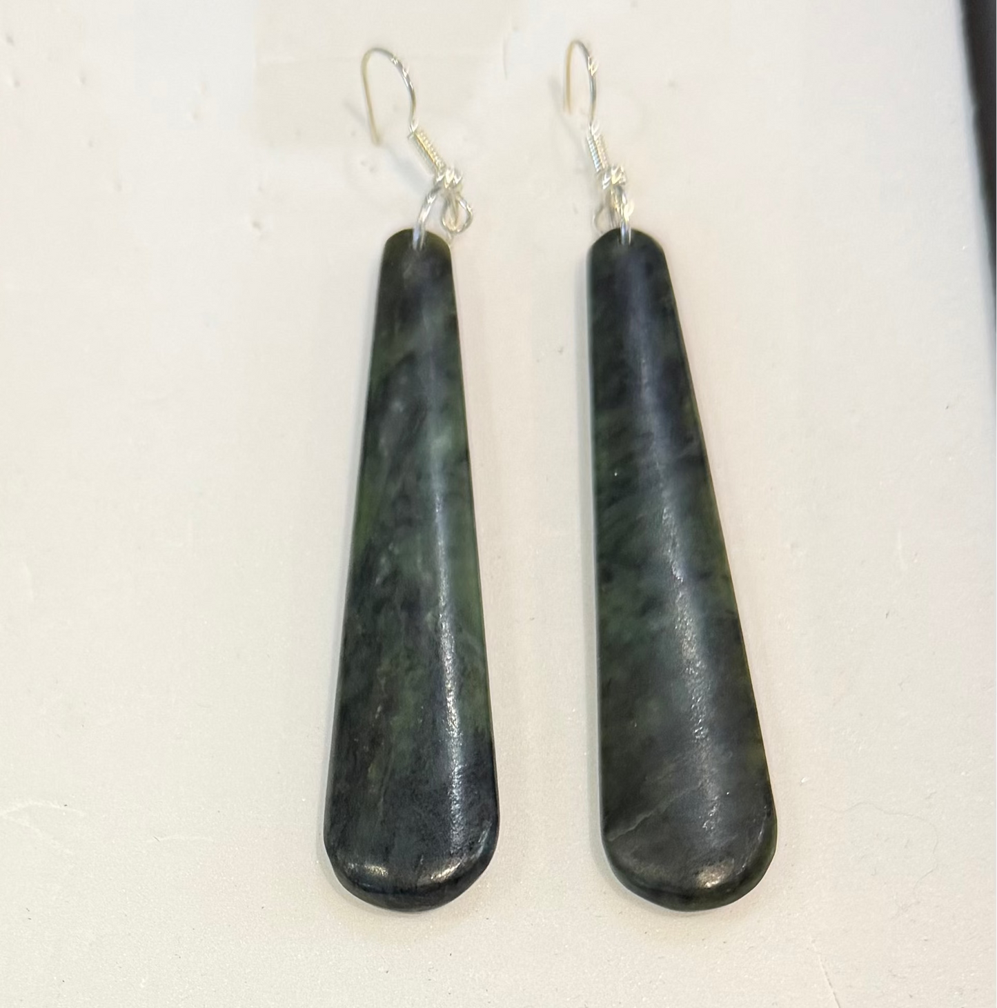 A pair of teardrop-shaped dark green and black stone drop earrings with silver fishhook wires, displayed on a white textured surface.