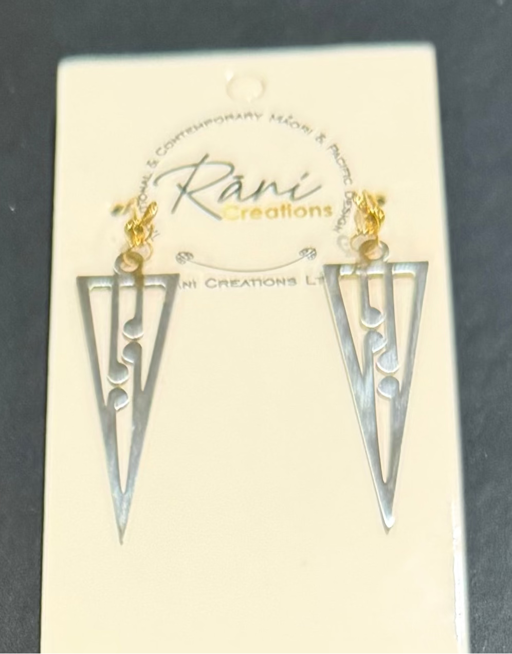 A pair of silver-toned geometric dangle earrings with gold-toned hooks, displayed on a cream-colored brand card that reads 'Rani Creations'. The earrings are photographed against a dark gray background.