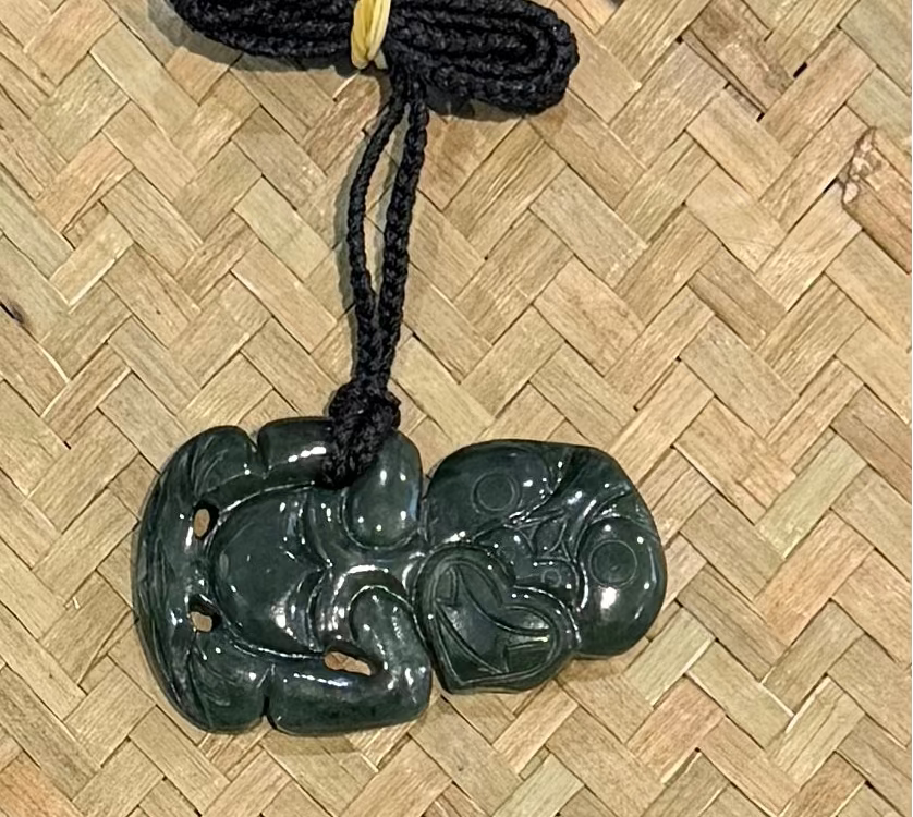 A dark greenstone carved jadeite pendant with a black braided cord, tied with a small yellow band, displayed on a woven straw mat background.