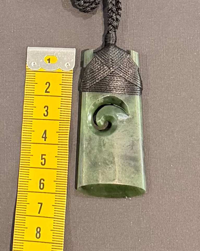 Greenstone pendant with spiral design on a string, next to a yellow ruler for scale.