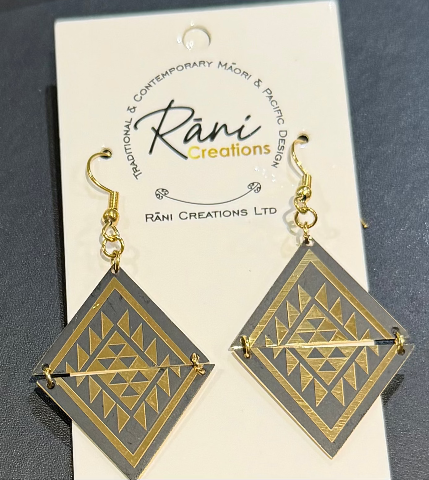 A pair of geometric drop earrings displayed on a white brand card. The earrings feature a dark gray stone inlay with a gold geometric pattern, suspended from gold hooks.