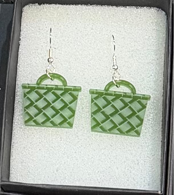 A pair of green woven basket-shaped dangle earrings displayed inside an open black gift box with white foam insert.
