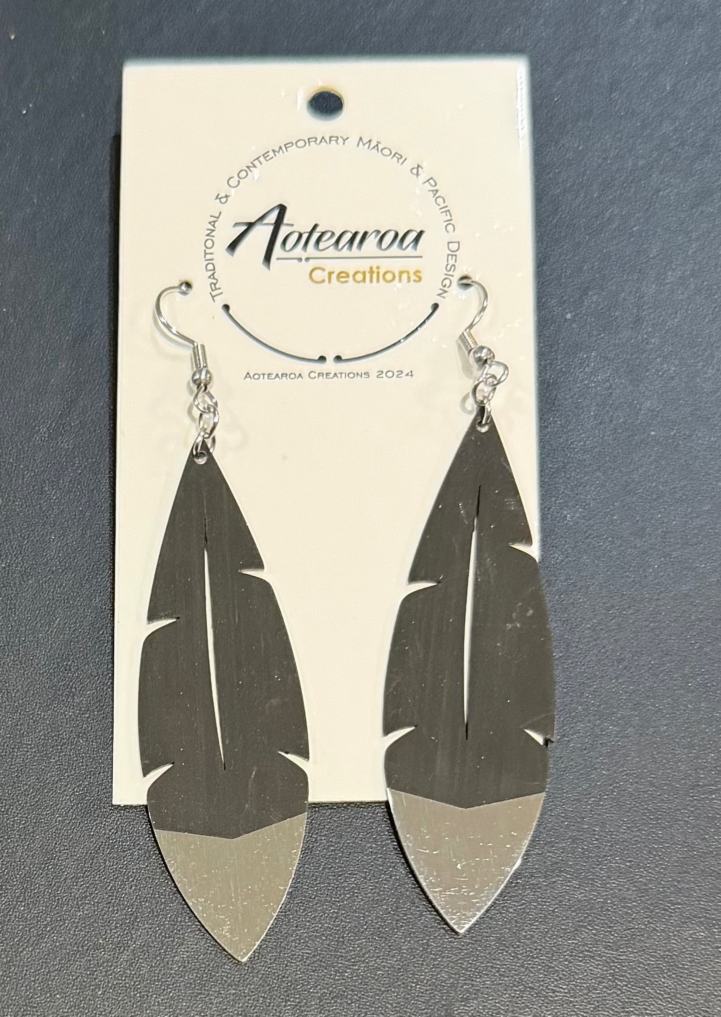 A pair of silver-toned dangle earrings shaped like stylized feathers, hanging from a white branded card that reads Aotearoa Creations, placed on a dark gray surface.