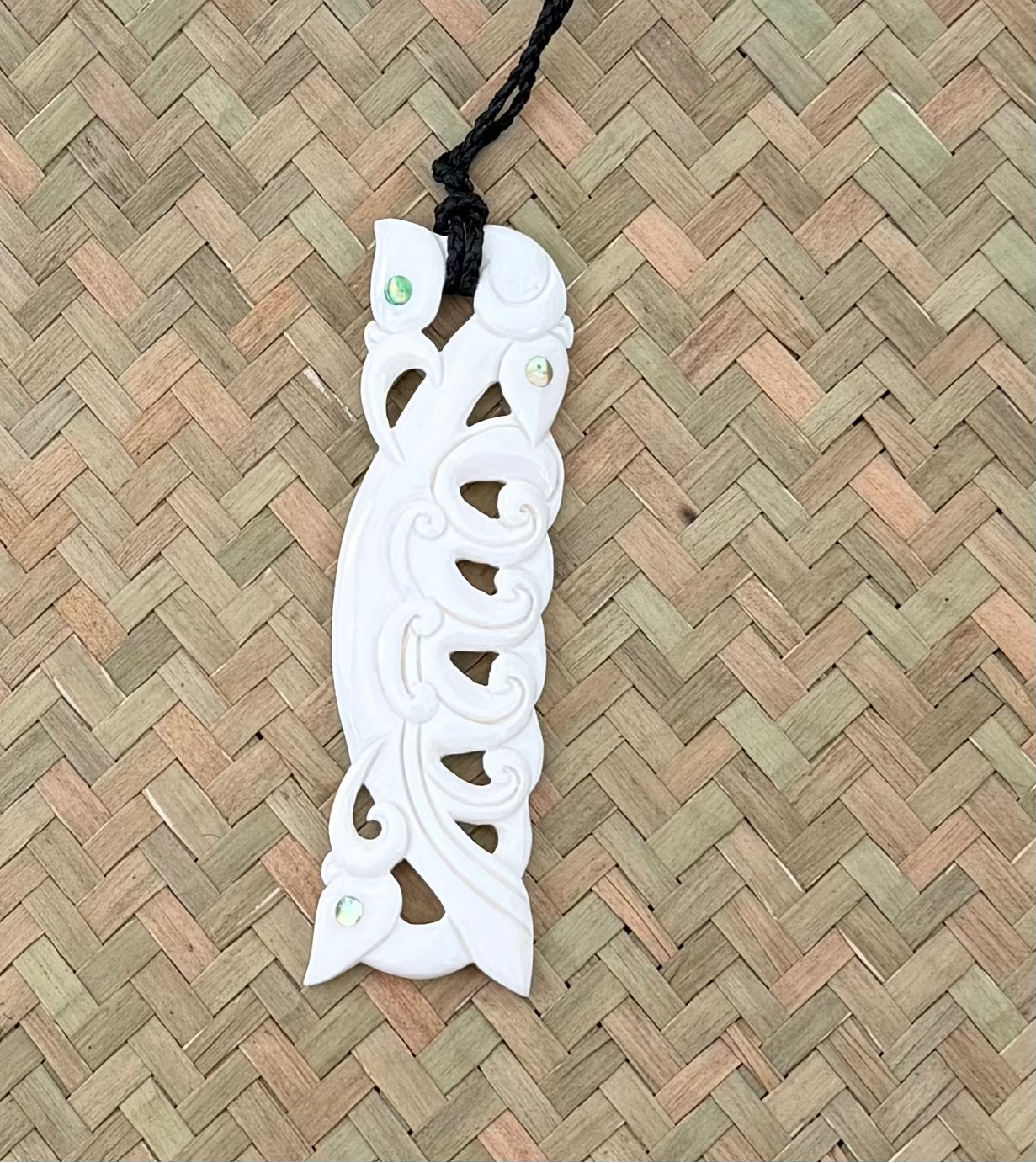 White manaia bone carved pendant on a black cord against a woven mat background
