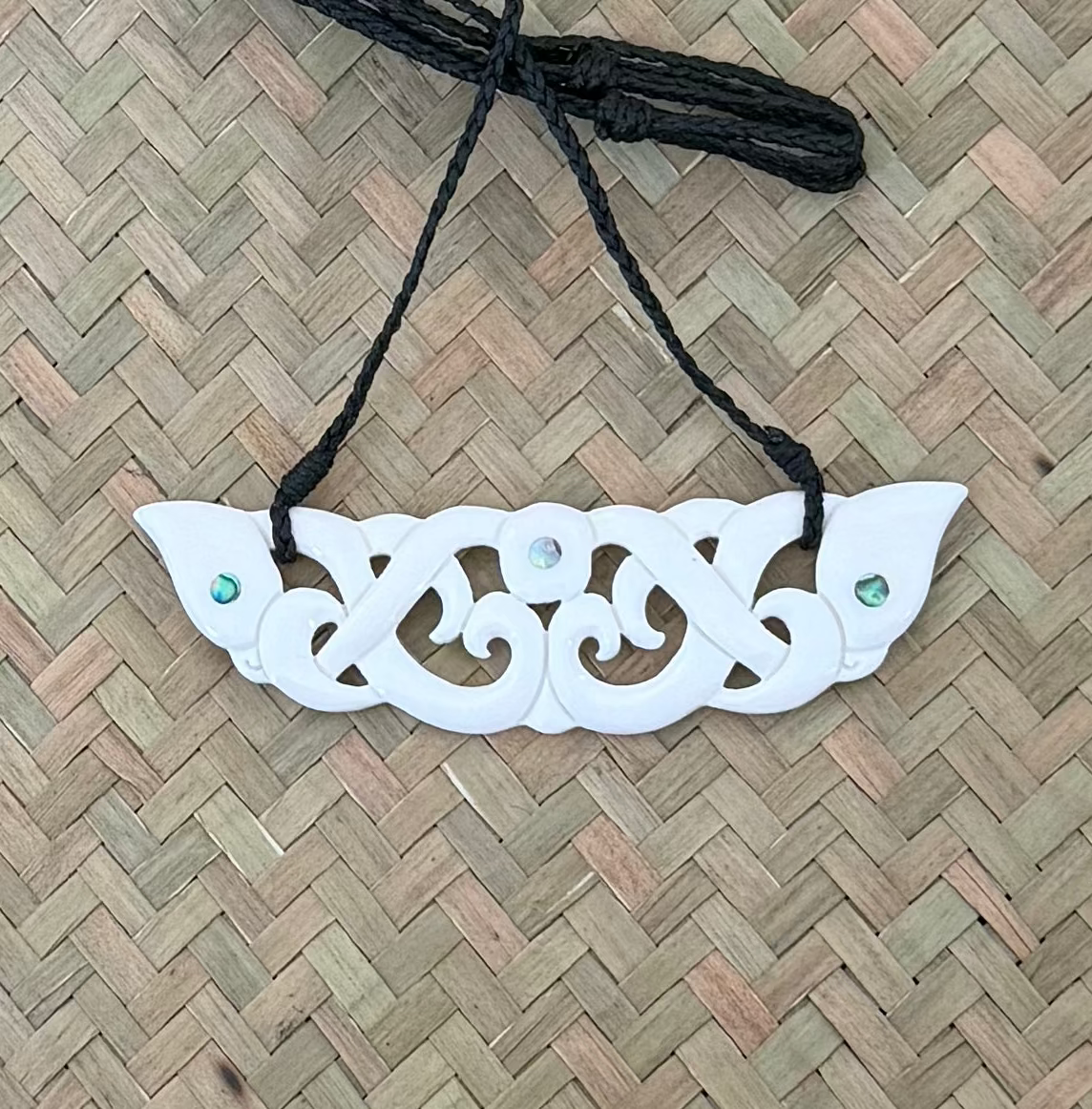 Decorative white MANAIA BONE pendant with green stones on a black cord against a woven background