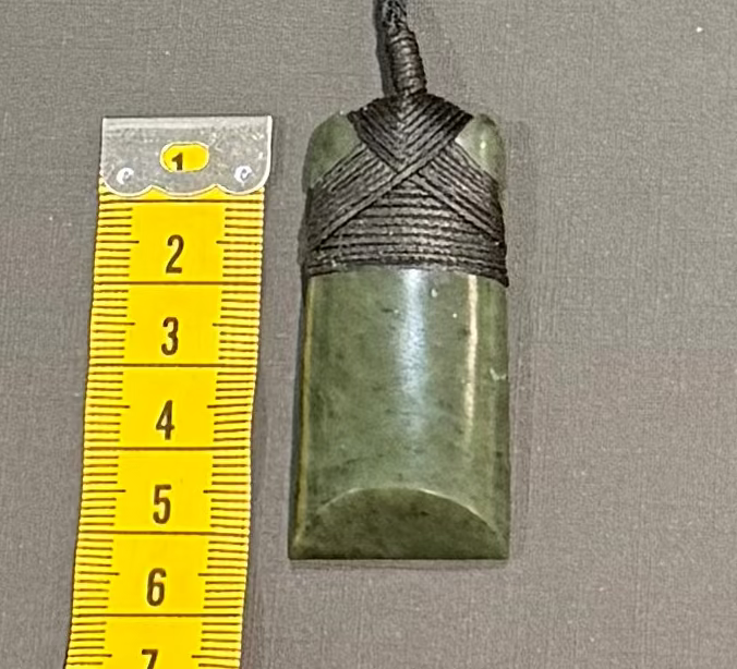 Green stone pendant with black cord on a gray background with a yellow measuring tape for scale.