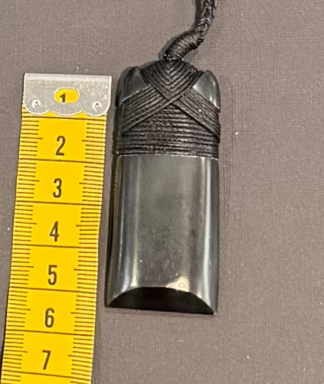 Black pounamu pendant on a string with a yellow measuring tape for scale on a gray background