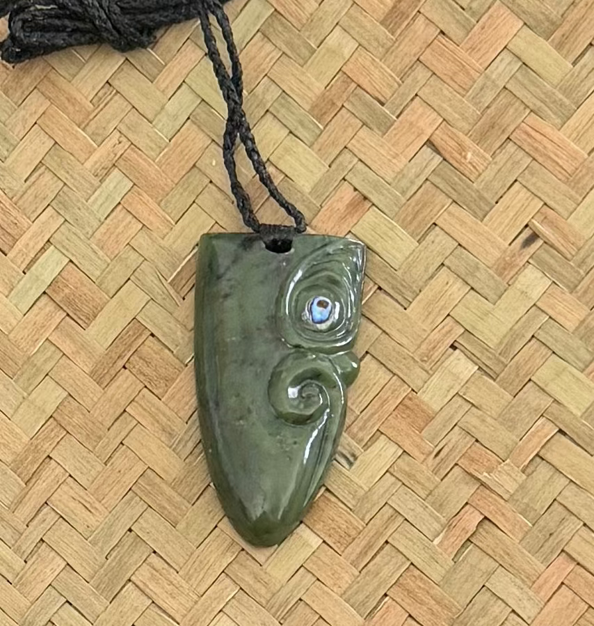 A carved greenstone pendant with a black cord, photographed on a woven tan straw mat background.