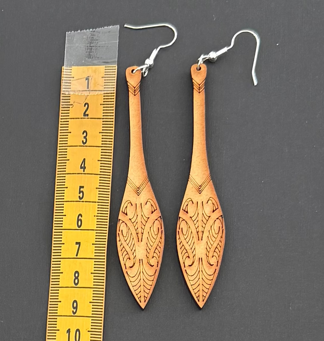 Pair of wooden Maori earrings with silver hooks on a dark surface with a ruler for scale.