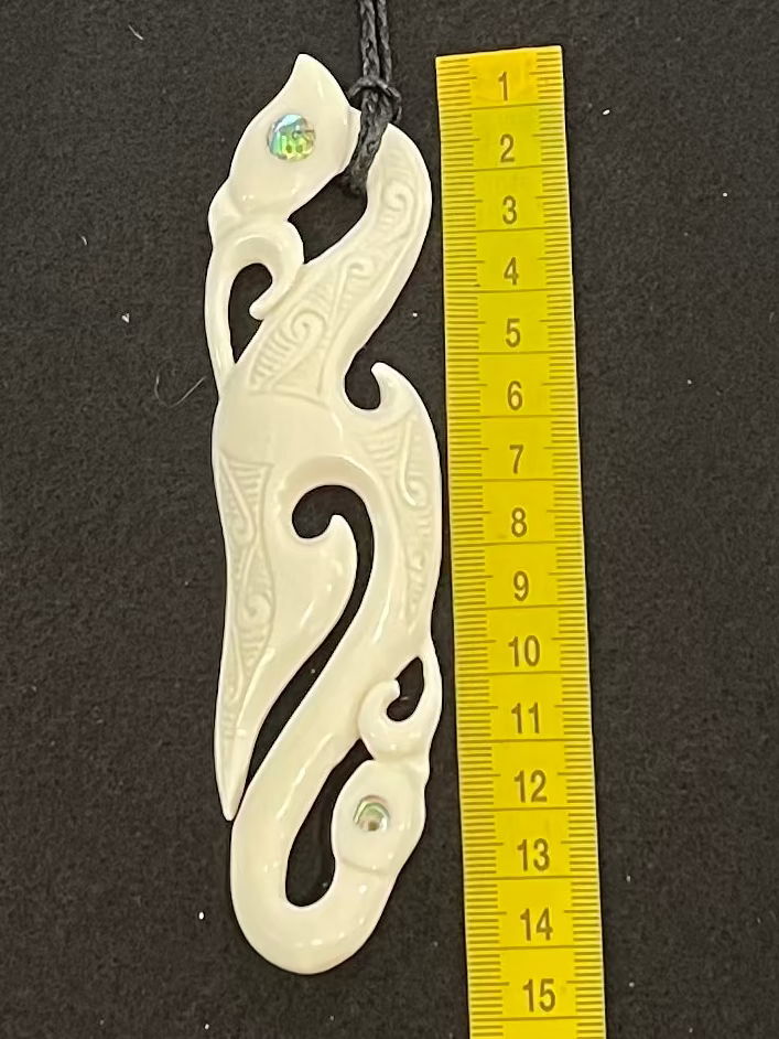 Carved bone manaia pendant next to a ruler on a black background