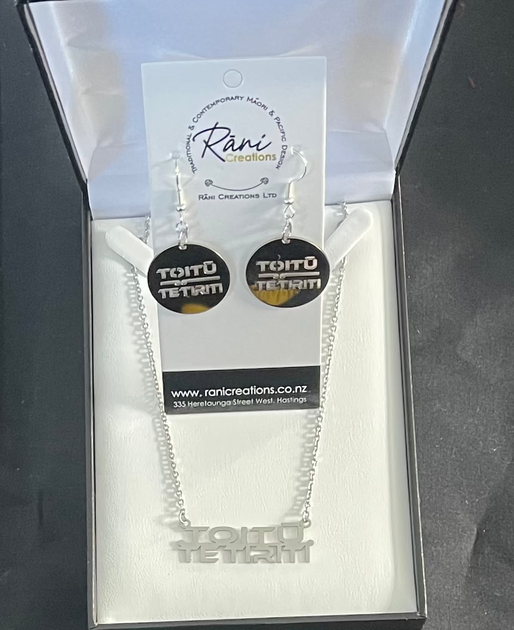 Silver necklace and earrings with 'Tortu Tetru' design in a Rani Creations packaging on a dark background.