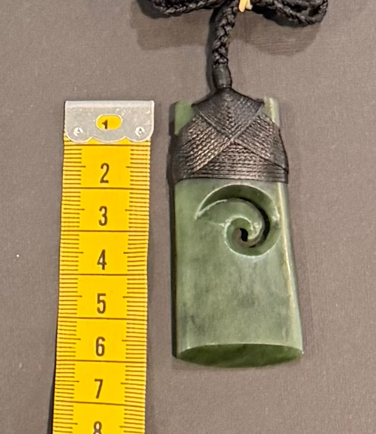 Greenstone pounamu carving on a cord with a yellow ruler for scale on a gray background