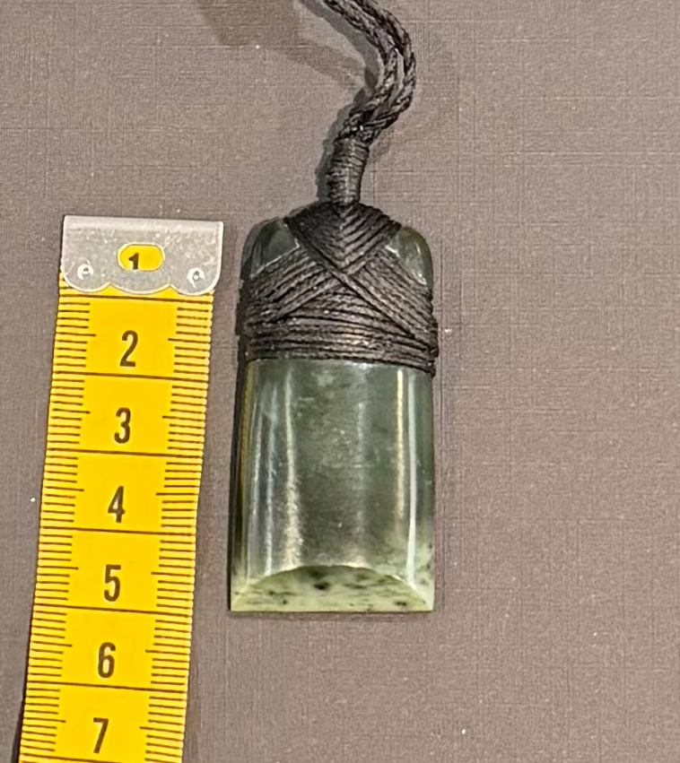 Greenstone pendant on a cord with a yellow measuring tape for scale on a gray background