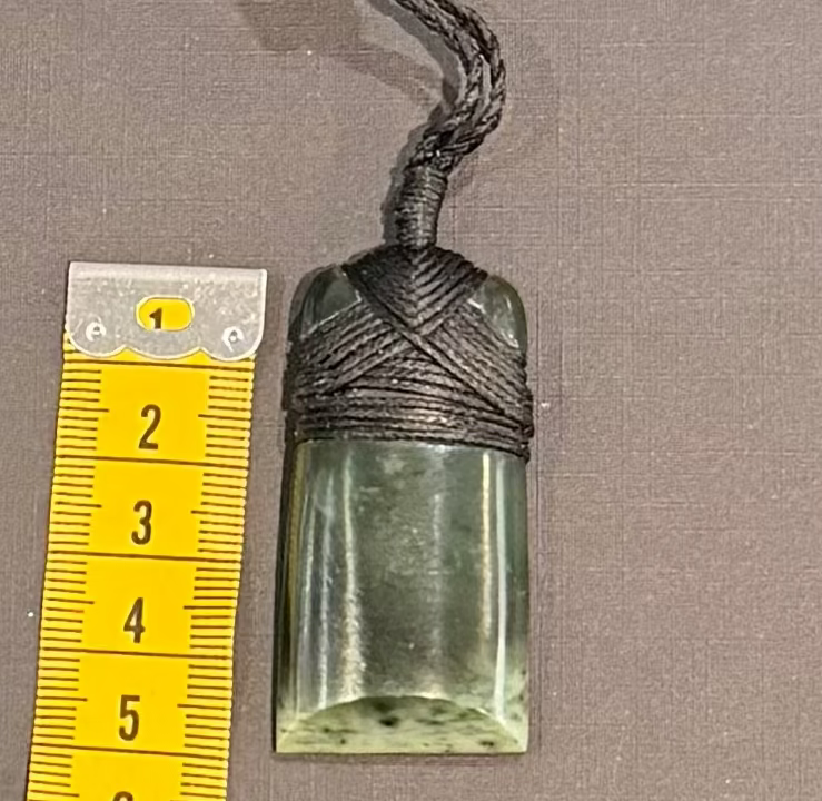 Greenstone pendant on a cord with a yellow measuring tape for scale on a gray background