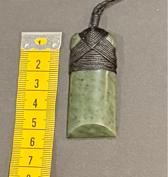 Greenstone pendant with cord next to a yellow measuring tape on a gray background