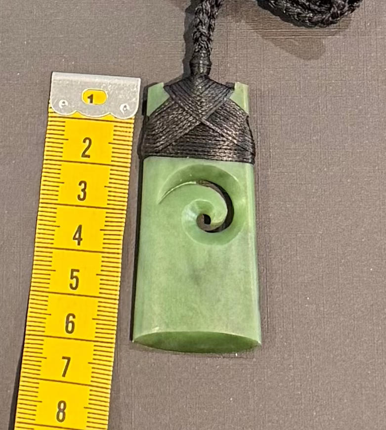 Greenstone jade pendant with spiral design on a brown background, next to a yellow measuring tape.