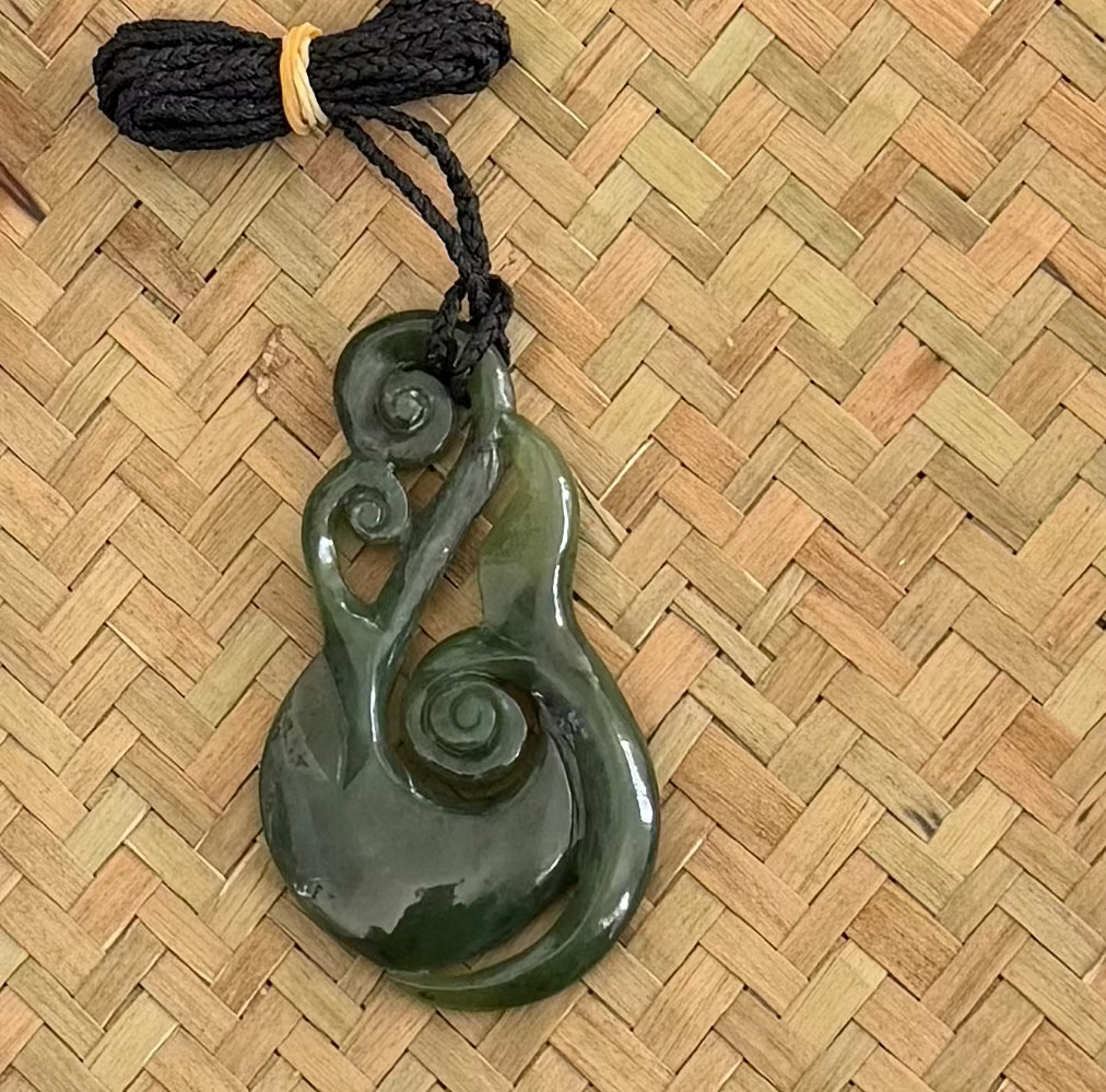 A carved dark green jade pendant with a South Pacific wai shape, attached to a black braided cord with a small yellow rubber band, displayed on a woven straw mat.