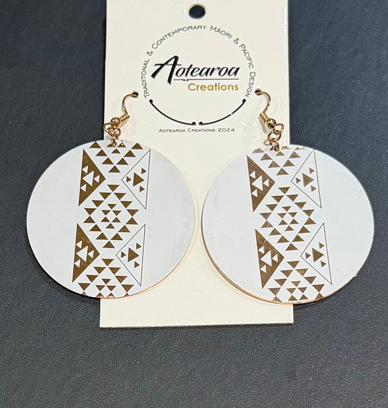 A pair of round white dangle earrings with a geometric gold triangle pattern, hanging from gold-toned hooks on a white paper display card. The card is branded 'Aotearoa Creations'.