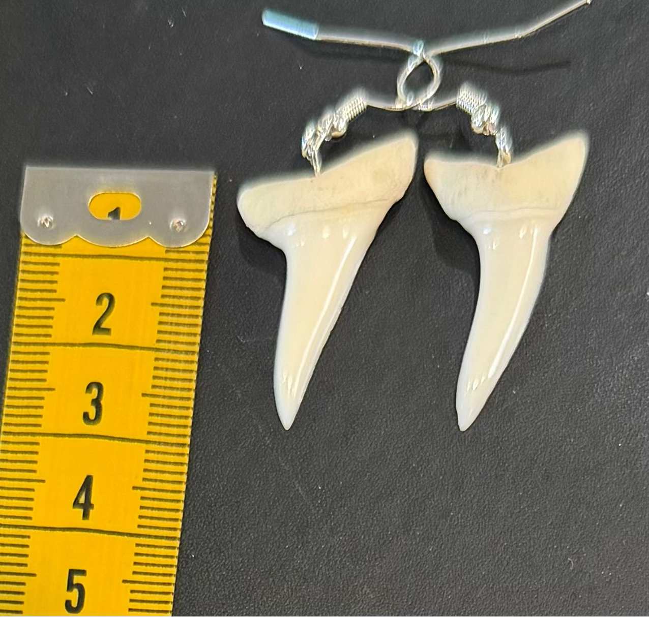 A pair of silver shark tooth drop earrings displayed next to a yellow measuring tape on a dark gray background.