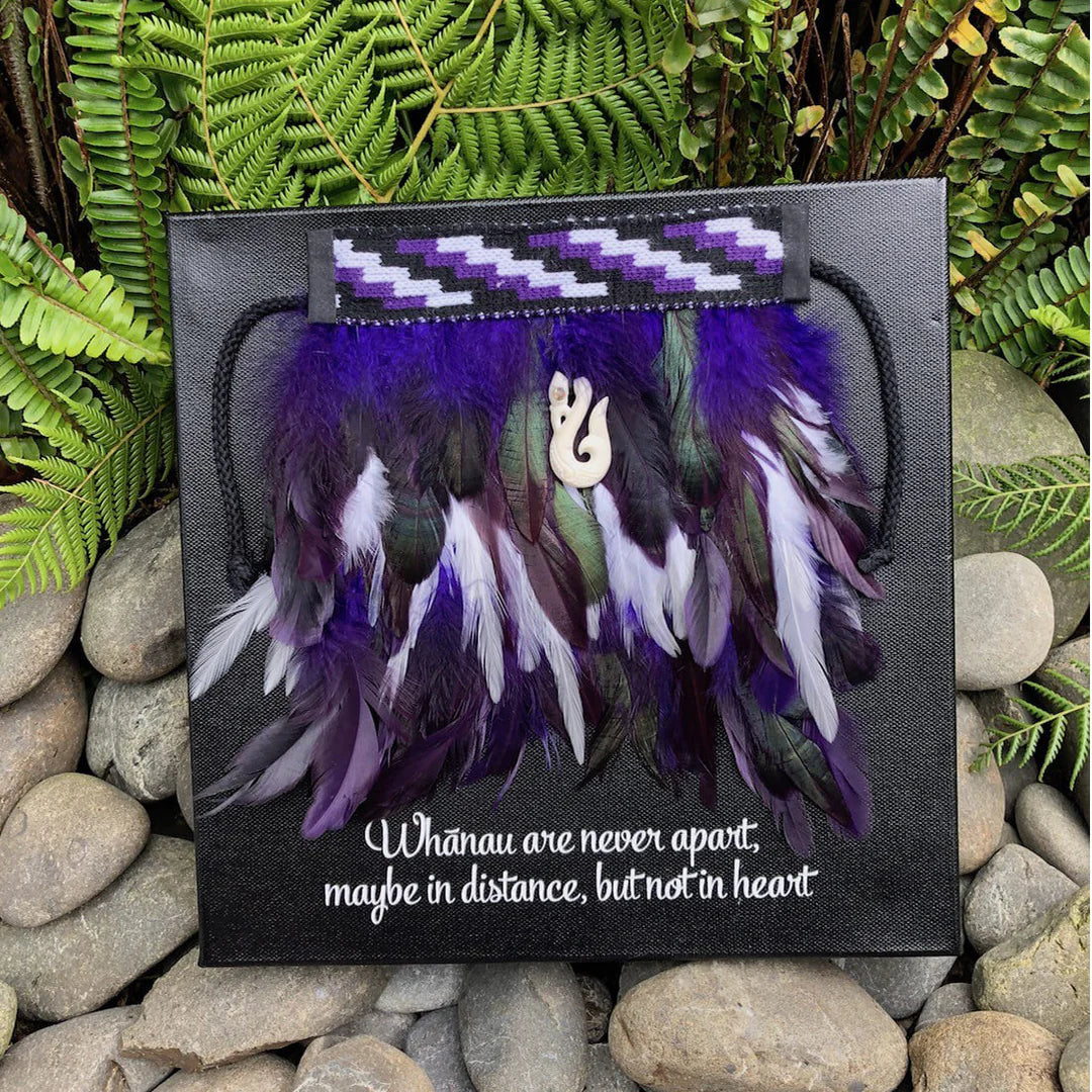 Decorative purple and white mini korowai on a black background, surrounded by green foliage and stones.