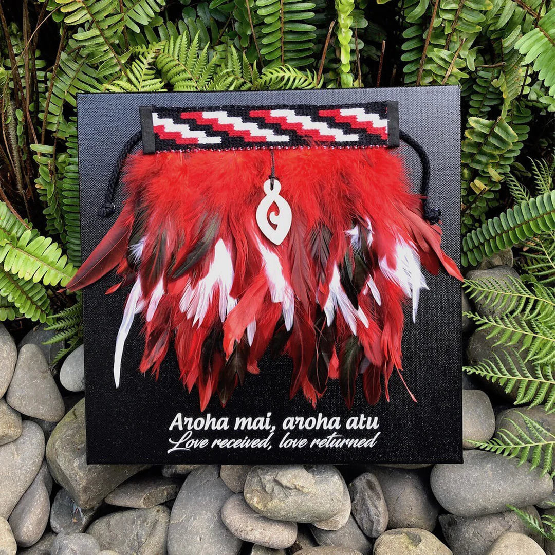Feathered red, white, and black korowai on canvas on a black background, surrounded by green ferns and stones.