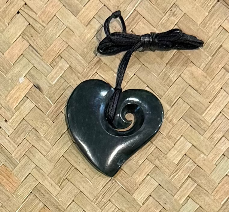 A greenstone heart-shaped pendant with a spiral cutout, attached to a black cord, placed on a woven straw mat background.