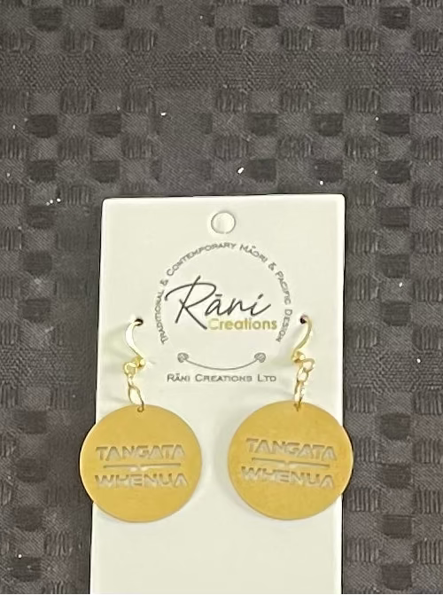 Gold tangata whenua earrings on a Rani Creations branded card against a textured gray background