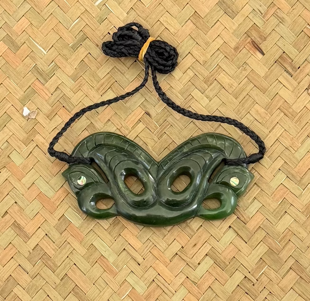 A dark green carved jade pendant with a black braided cord, displayed on a woven straw mat background.