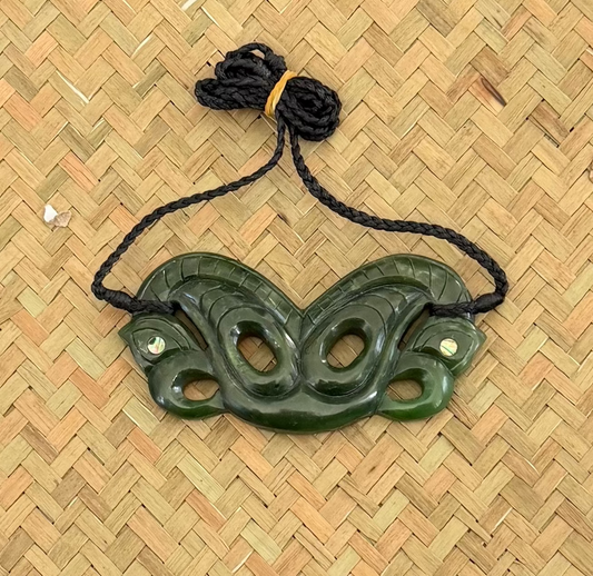 A dark green carved jade pendant with a black braided cord, displayed on a woven straw mat background.