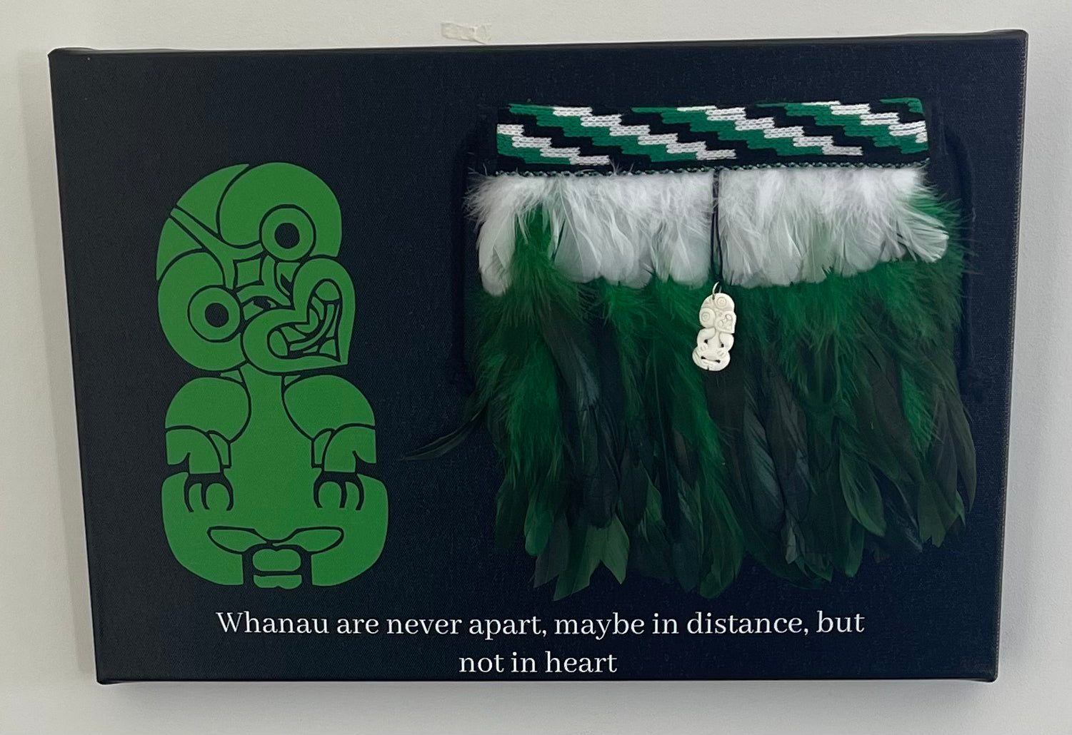 Green Feathered Maori korowai with green and white design on a black box with a green tiki figure and text.
