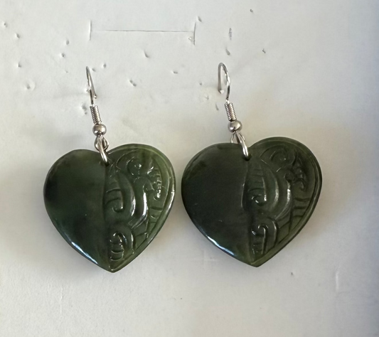 A pair of heart-shaped dark green carved stone pendants with silver fishhook ear wires, displayed on a textured off-white background.