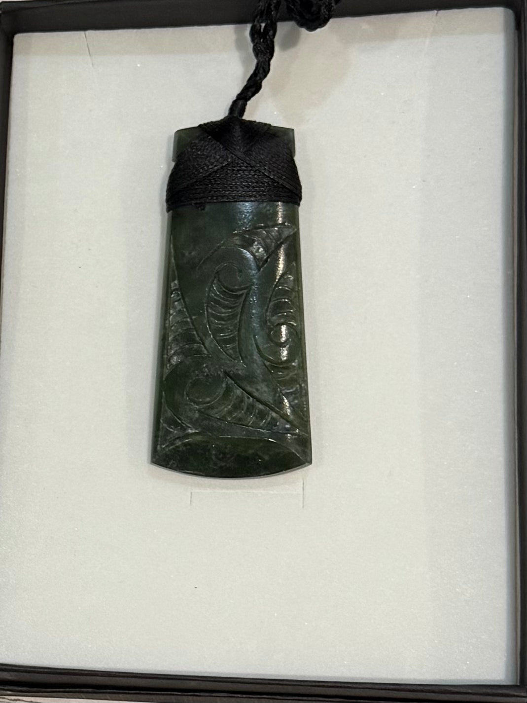 Carved greenstone pendant on a black cord displayed in a box with white tissue paper.