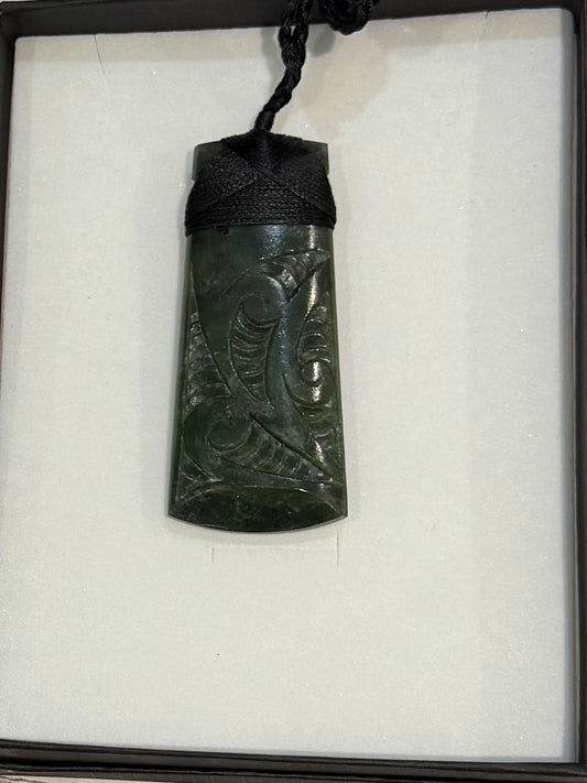 Carved greenstone pendant on a black cord displayed in a box with white tissue paper.