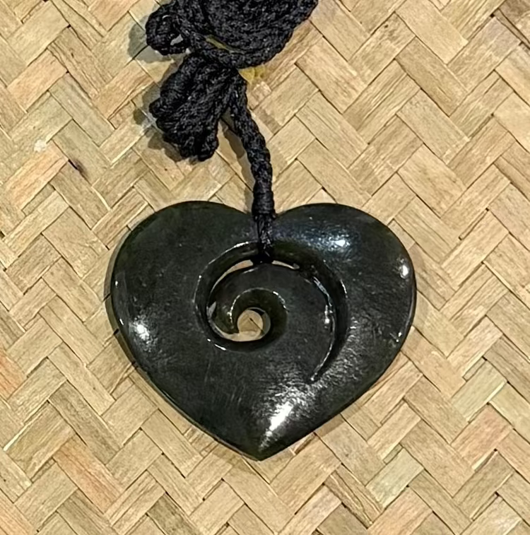A greenstone carved heart-shaped pendant with a spiral cutout, attached to a black braided cord, displayed on a woven straw mat background.