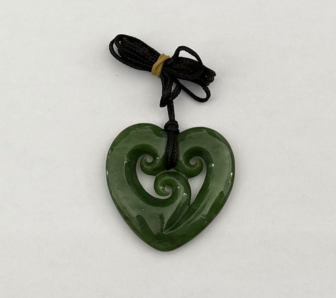 A green carved heart-shaped pendant with a swirl motif, attached to a black cord with a small tan tie, shown on a plain light background.