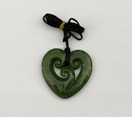 A green carved heart-shaped pendant with a swirl motif, attached to a black cord with a small tan tie, shown on a plain light background.